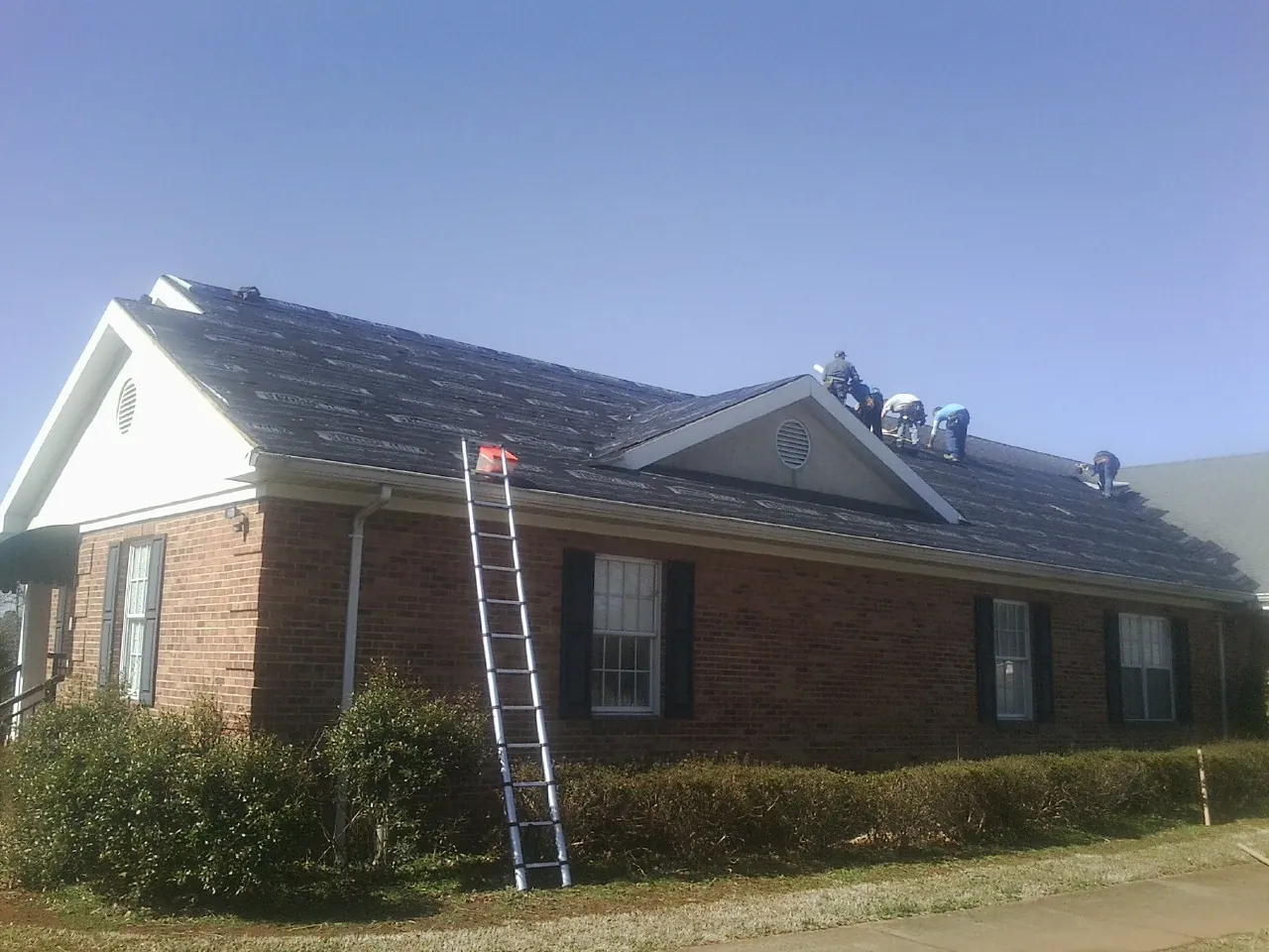 Crew tearing shingles off a brick ranch roof with a ladder set — Emergency Roof Repair work by our crew in Cogswell