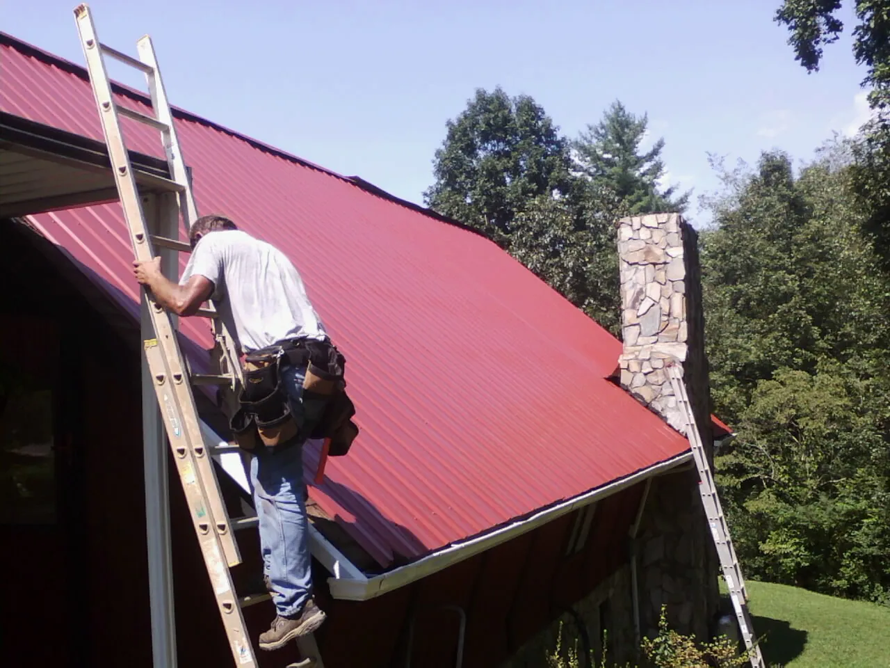 Roofer on a ladder beside a red metal roof and stone chimney — Soffit & Fascia Repair by Roofing Brothers in Cogswell
